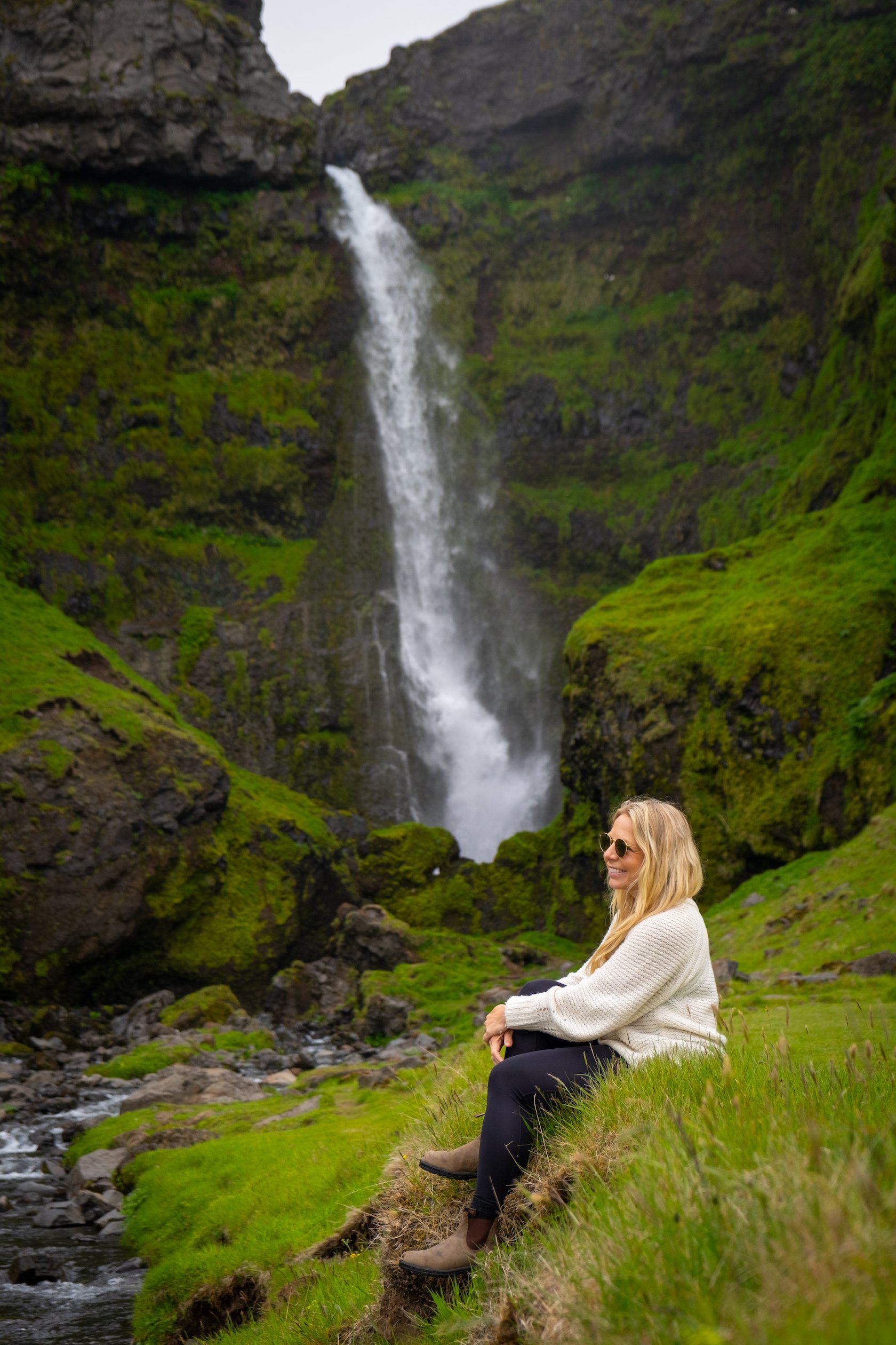 Natasha Sits Along A Grassy Stream With A Waterfall Behind Her In Iceland