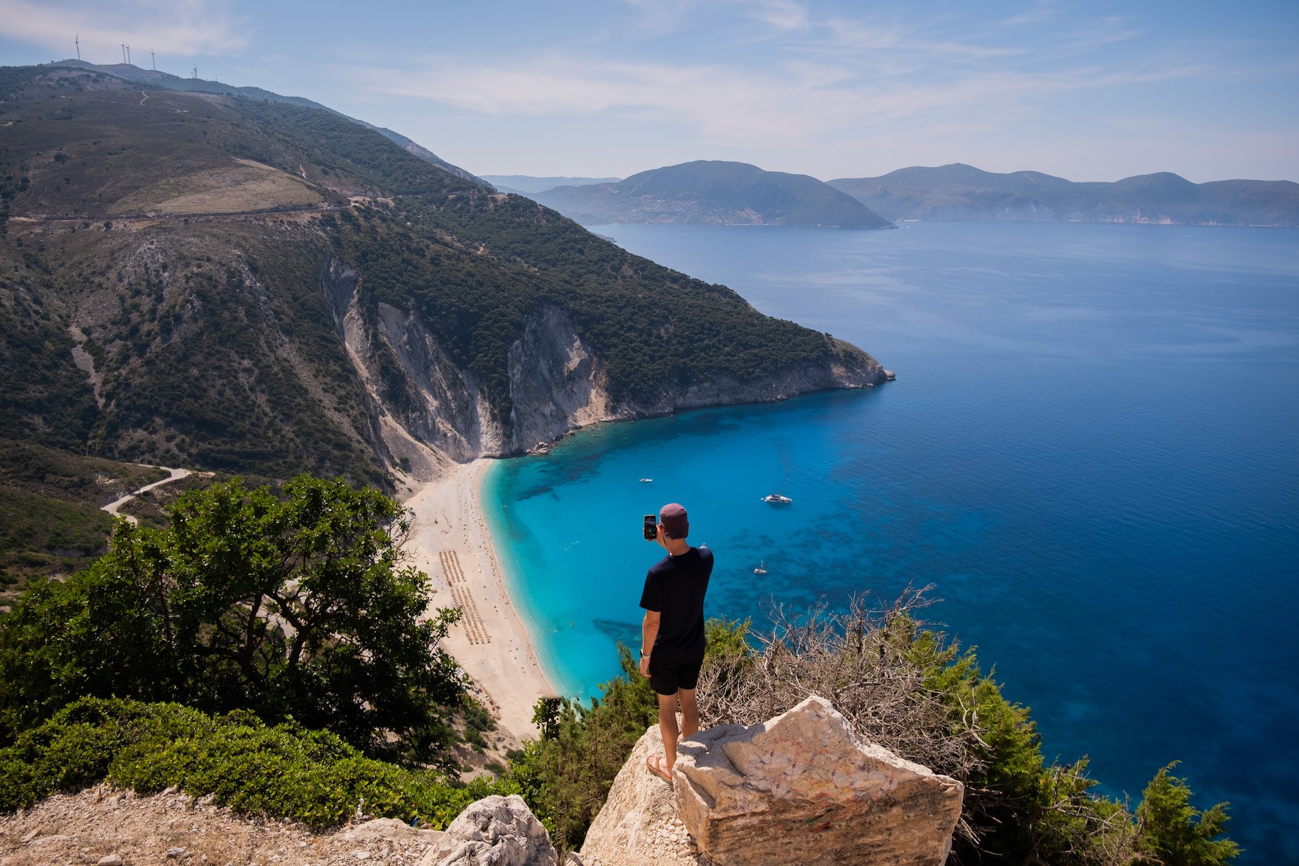 Cameron Stands Above Myrtos Beach Viewpoint With Phone Taking A Photo