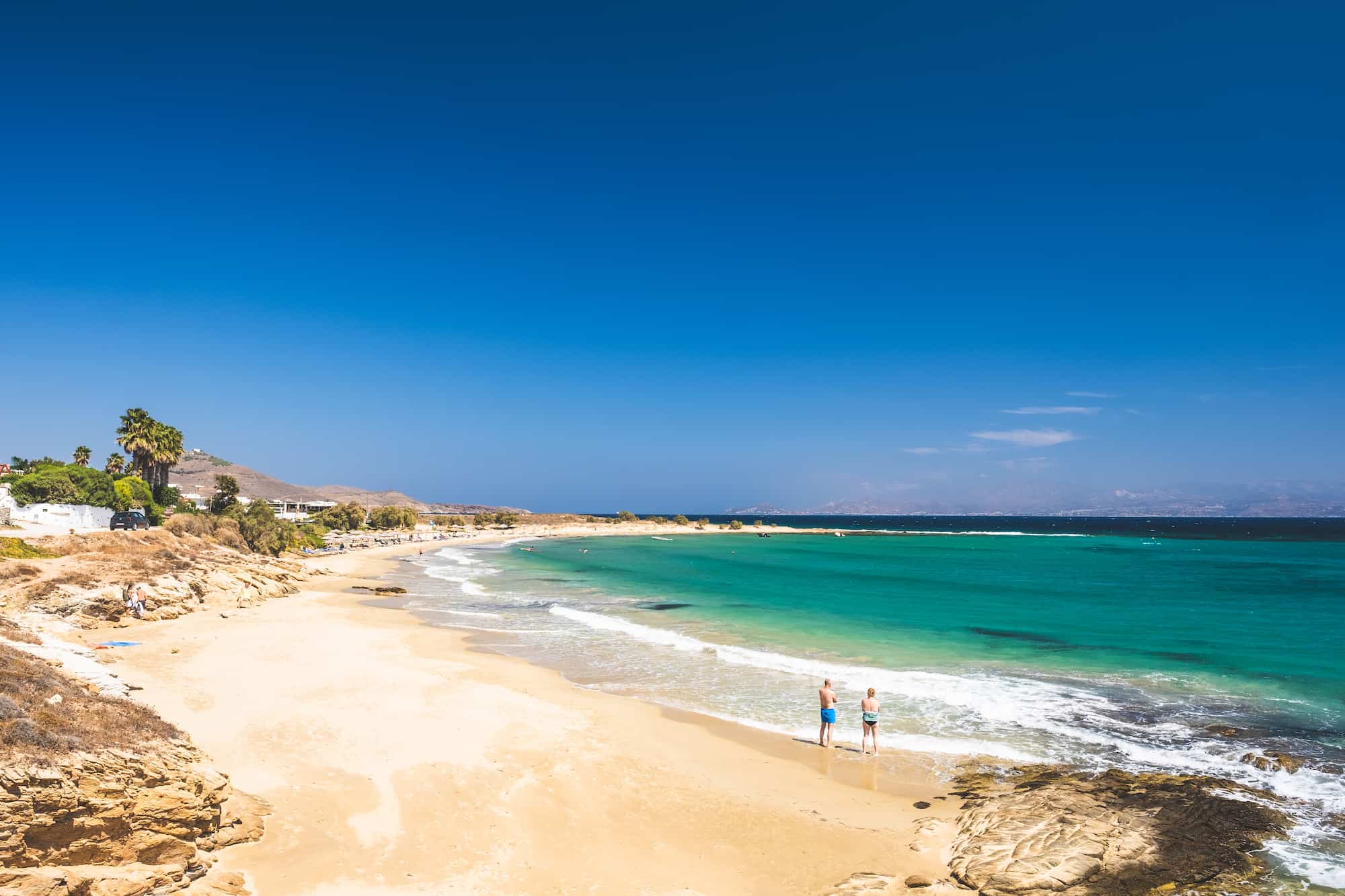 Two People Stand On The Water Edge At Golden Beach On Paros